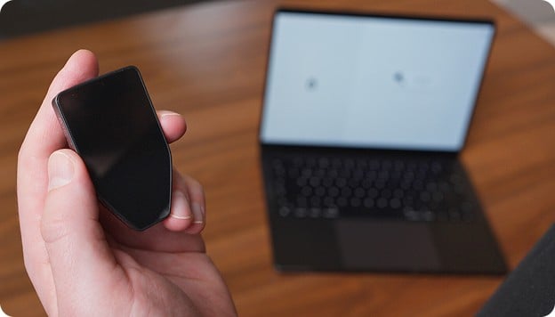 Hand holding a small black device in front of an open laptop on a wooden desk.