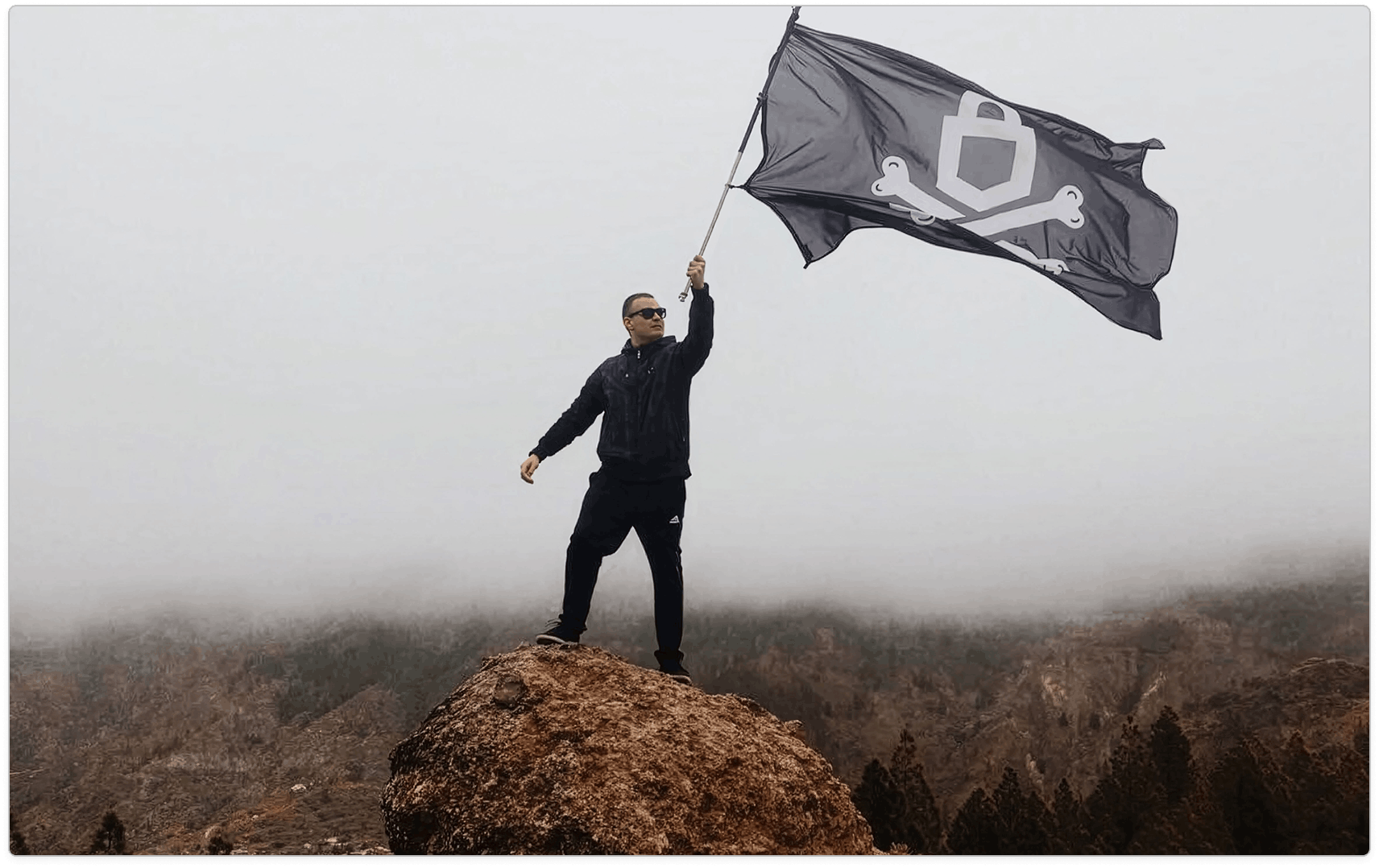 Person in black standing on a rocky summit and holding a large black emblem flag above a hazy forest.