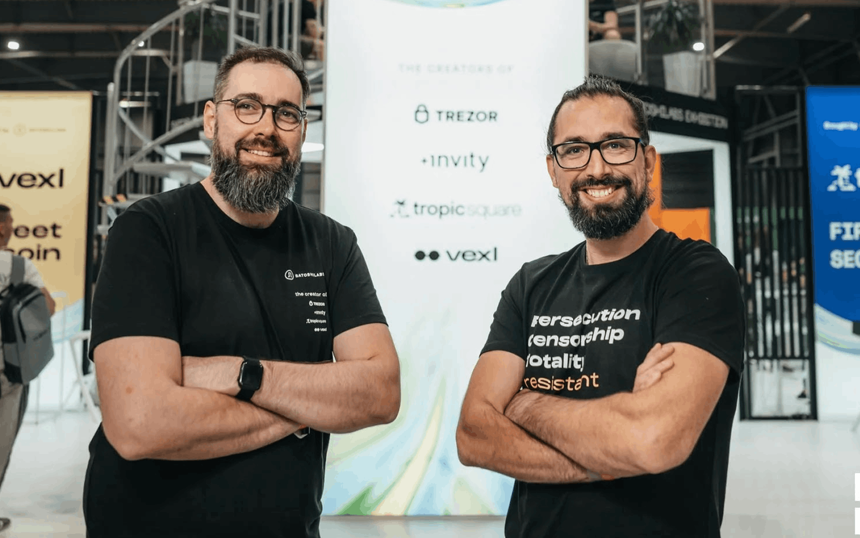 Two bearded men in black T-shirts pose with crossed arms at a technology conference.
