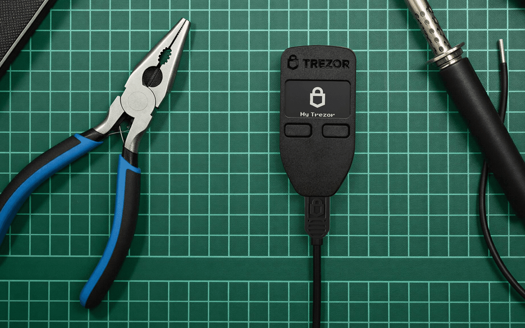 Trezor hardware wallet with a lock screen, pliers, and soldering tools on a green gridded cutting mat.